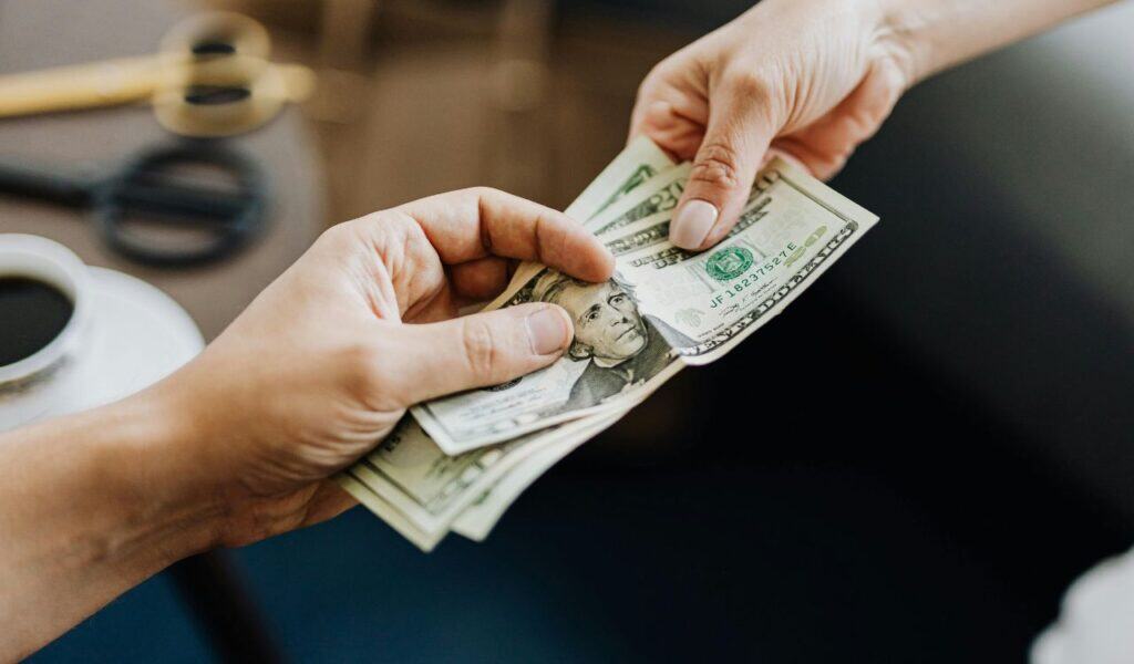 A close-up of hands exchanging U.S. dollar bills, representing an in-person cash transaction and emphasizing the importance of inspecting items before payment to avoid upfront payment scams.
