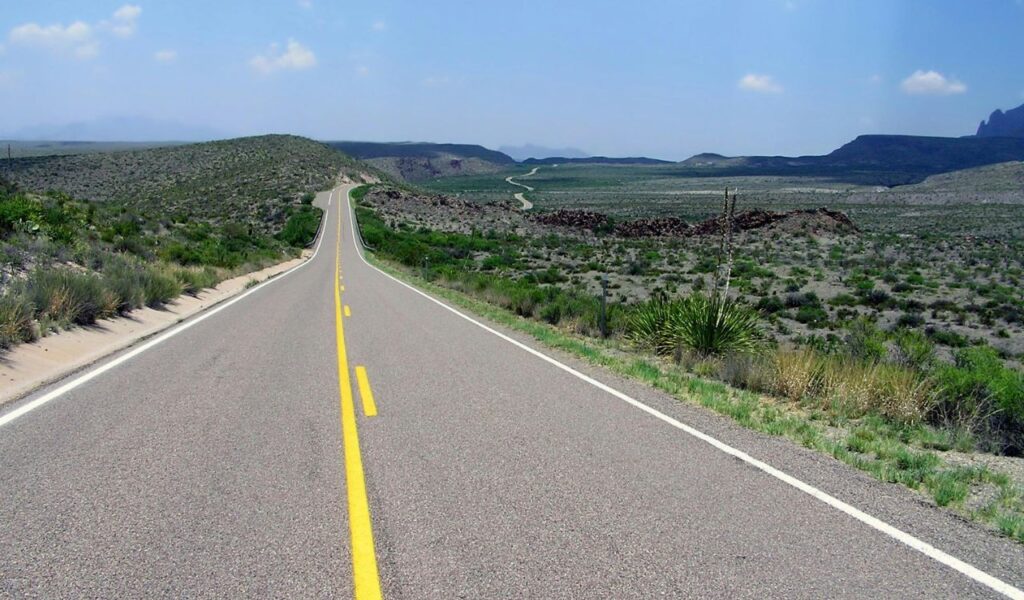  Long, empty desert highway with a double yellow line stretching into the distance through arid, scrub-covered terrain between New Mexico and Texas under a clear blue sky.