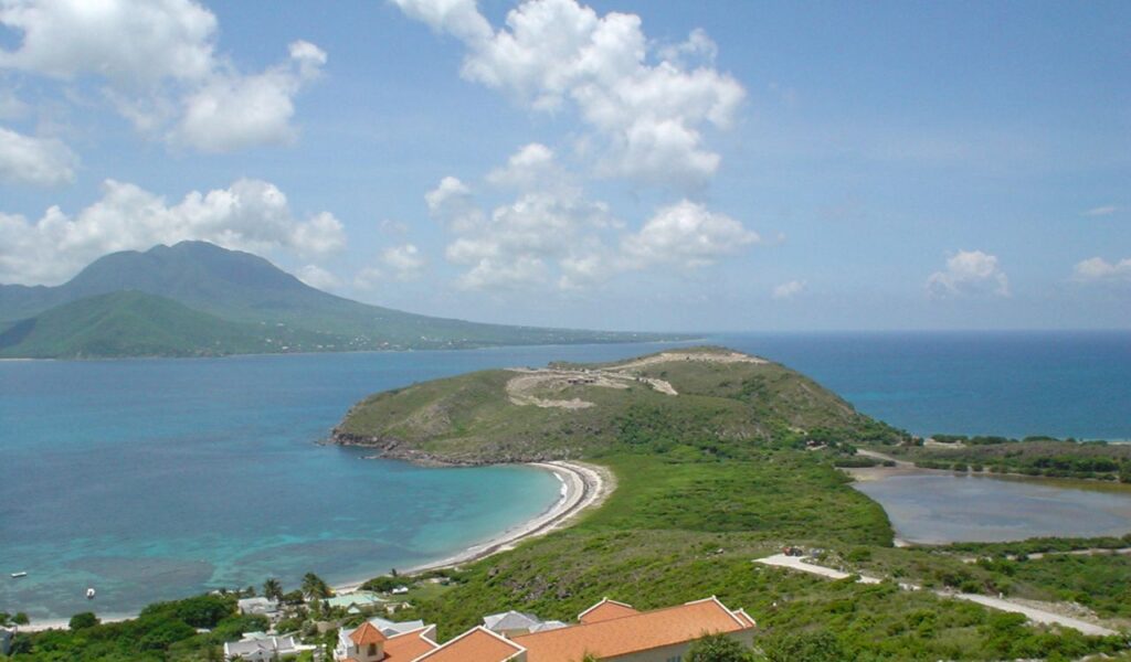 Scenic view of a tropical coastline with clear turquoise waters, a lush green hill, and a distant mountain under a bright sky