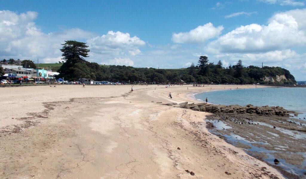Sunny beach in New Zealand with a sandy shoreline, calm waters, and people enjoying the beautiful coastal scenery.