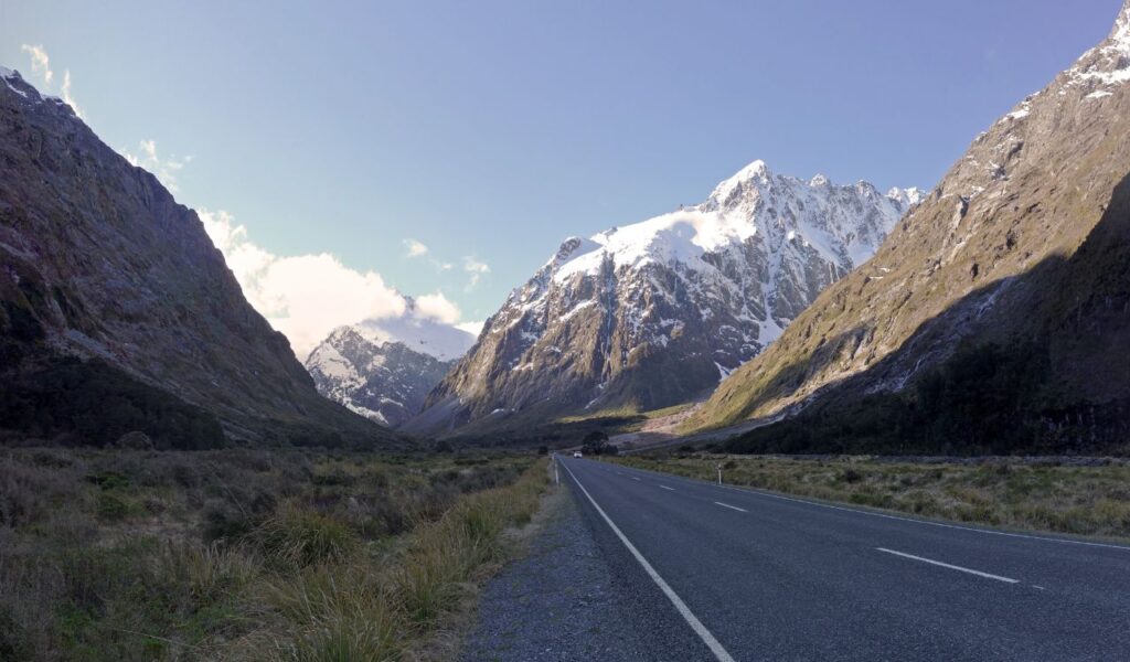 Empty road cutting through Fiordland National Park with snow-capped mountains under a clear sky in New Zealand.