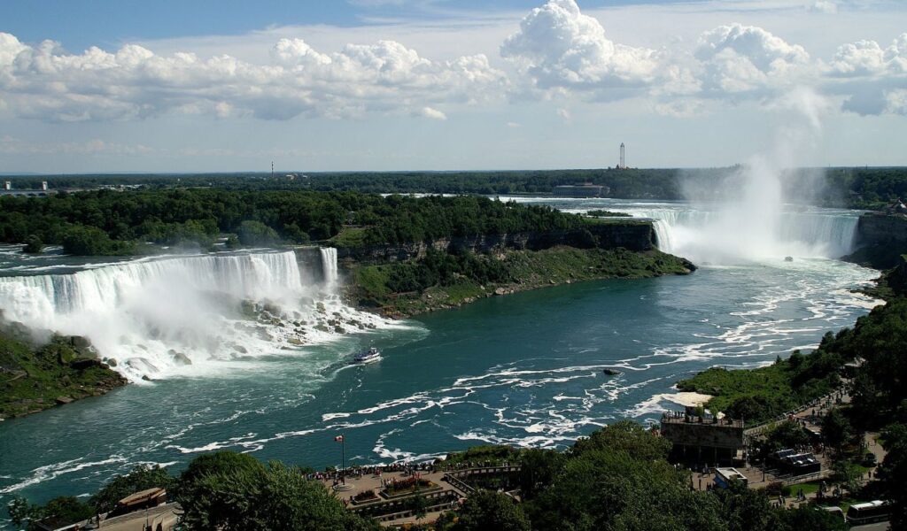 A wide view of Niagara Falls showing both the American and Horseshoe Falls, with mist rising and a tour boat approaching the base, surrounded by lush greenery and crowds of visitors on both sides.
