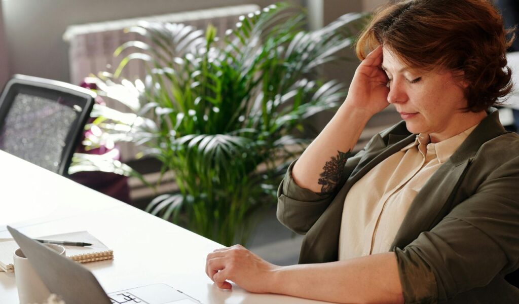 A woman sits at a desk with her eyes closed and hand on her temple, appearing stressed or fatigued, with a laptop and notebook in front of her.