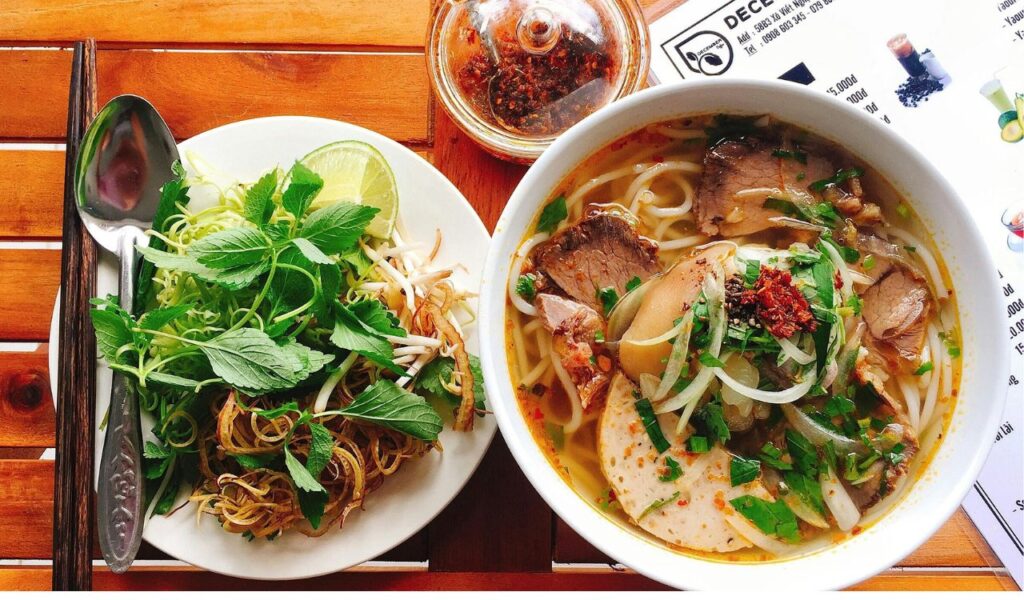 A bowl of Vietnamese noodle soup with sliced meat, herbs, and vegetables, served alongside a plate of fresh garnishes and utensils, placed on a wooden table with a partially visible menu.