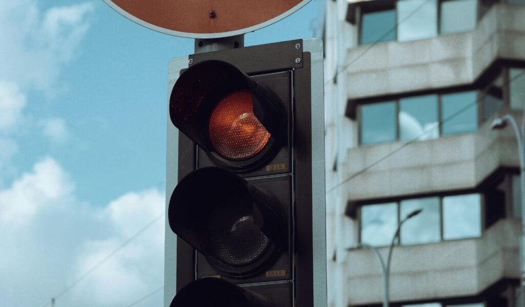 Close-up of a red traffic light with a “No Right Turn on Red” sign above it, set against a backdrop of a city building and cloudy sky.