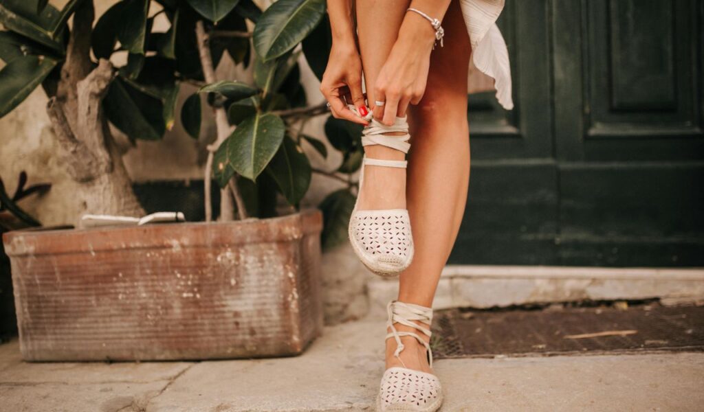 A close-up of a person tying their white lace-up shoes, standing in front of a plant with a green door in the background.