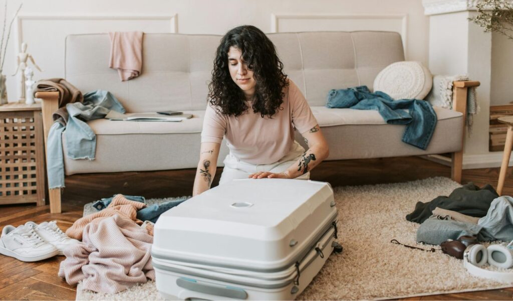 Young woman kneeling on the floor while packing a white suitcase, surrounded by clothes and travel essentials in a cozy living room with a sofa and wooden floor.