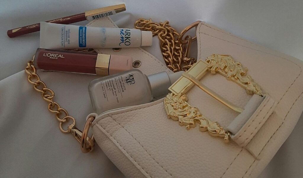 A beige handbag with a gold chain and clasp lying on a surface, surrounded by cosmetics including lip gloss, lip liner, face serum, and a small tube of cream.