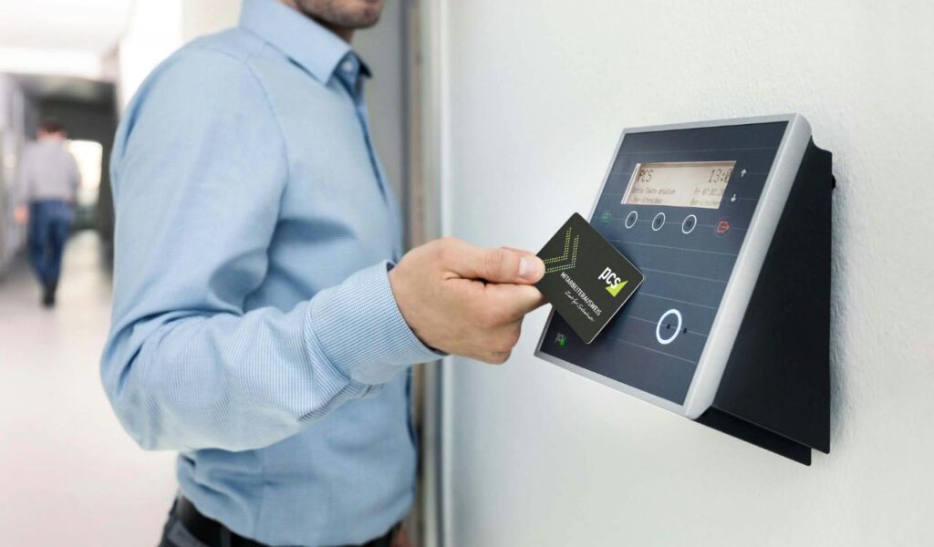 Man in a blue shirt holding an RFID card near a card reader terminal on a wall, preparing to scan for access.