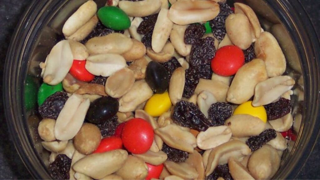 Close-up of a trail mix containing roasted peanuts, raisins, and colorful candy-coated chocolate pieces in a round container.