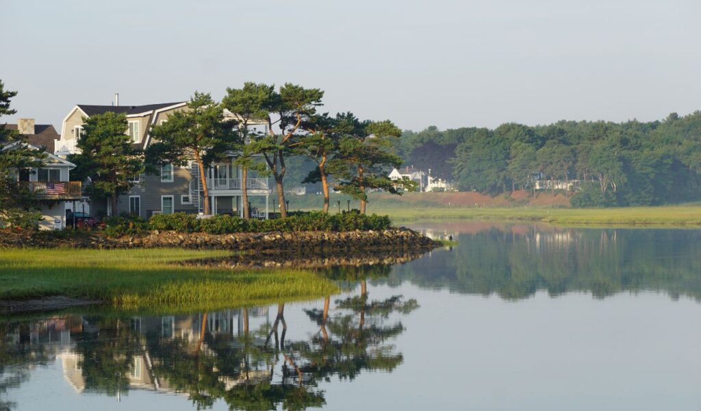 Waterfront homes and pine trees reflected in a calm river in Ogunquit, Maine, with misty forested hills in the background.