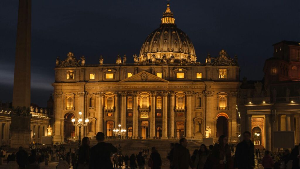St. Peter's Basilica illuminated at night with its grand dome and detailed façade, surrounded by visitors in St. Peter's Square.