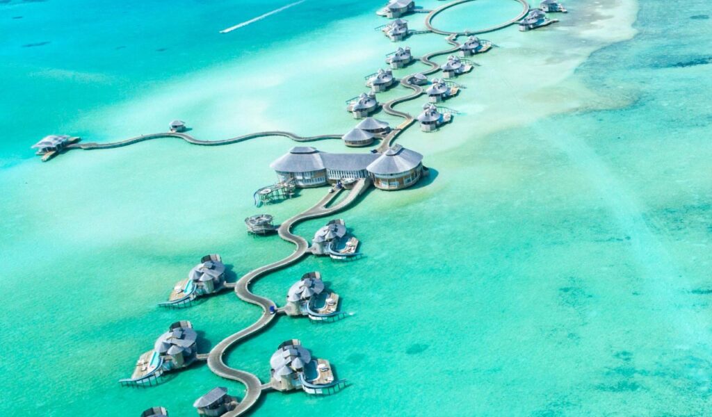 Aerial view of luxury overwater villas connected by winding wooden walkways across turquoise blue waters in a remote tropical paradise.