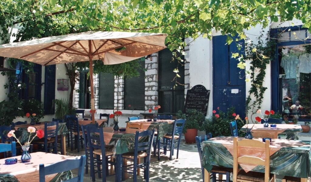 Charming Greek taverna with blue chairs and shaded outdoor seating under leafy vines and a large umbrella, perfect for alfresco dining.