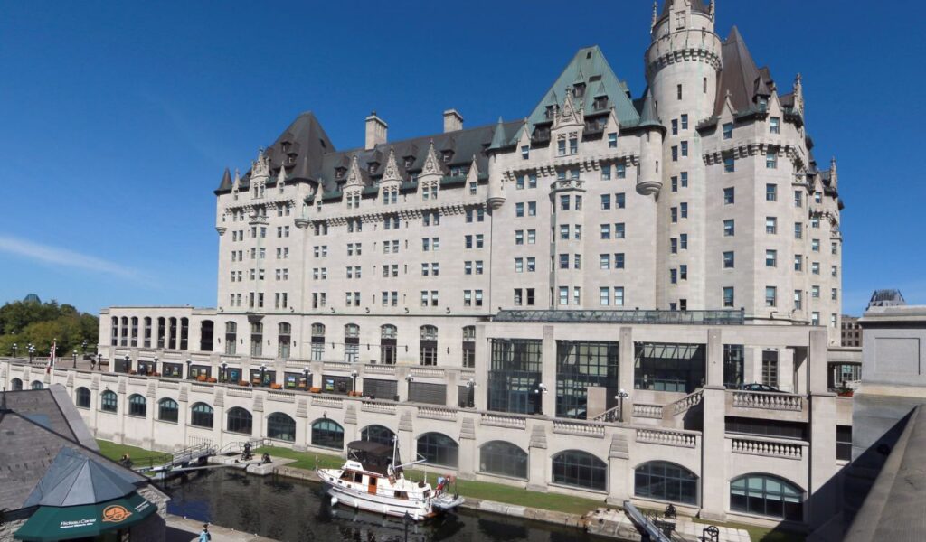 The historic Fairmont Château Laurier hotel in Ottawa, Ontario, overlooking the Rideau Canal with boats docked along the waterway.