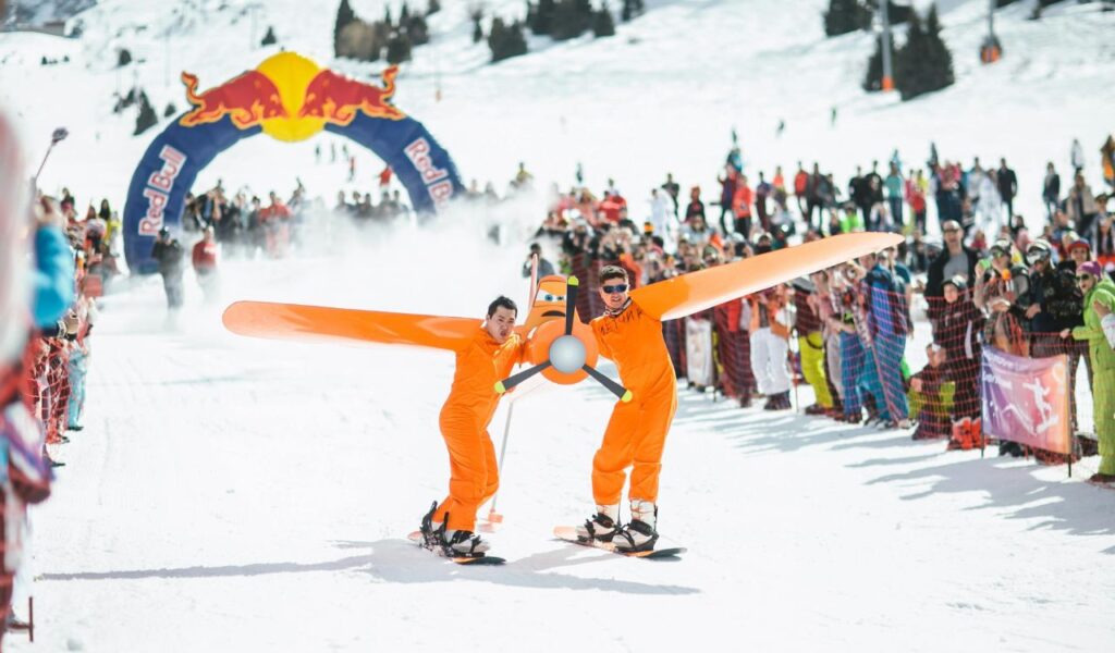 Red Bull Flugtag event in a snowy mountainous area with participants dressed in orange outfits, carrying a large airplane prop while snowboarding.