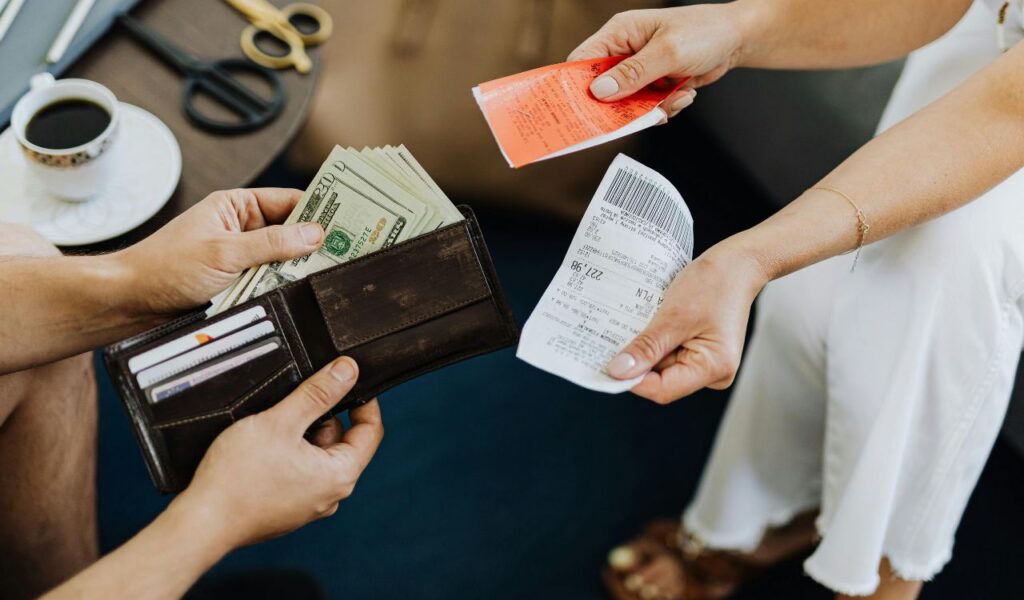 A person holds a wallet full of cash while another hands over receipts, symbolizing payment and expenses in a close-up transaction scene.