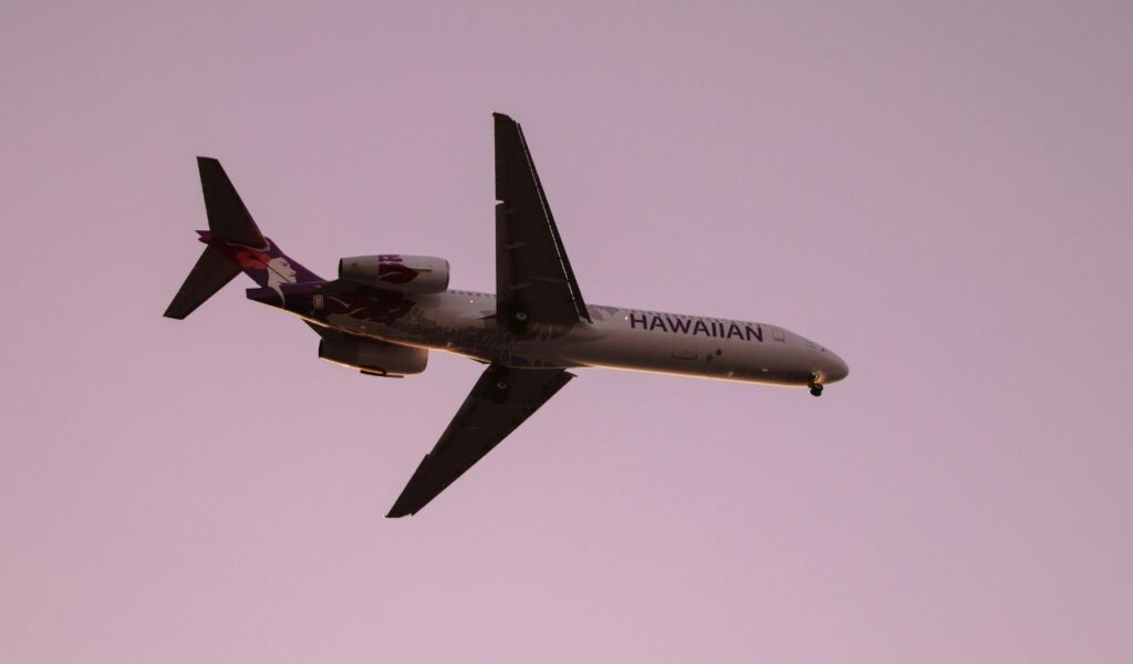 A Hawaiian Airlines jet soars through the sky at sunset, silhouetted against a soft pink backdrop during its flight.