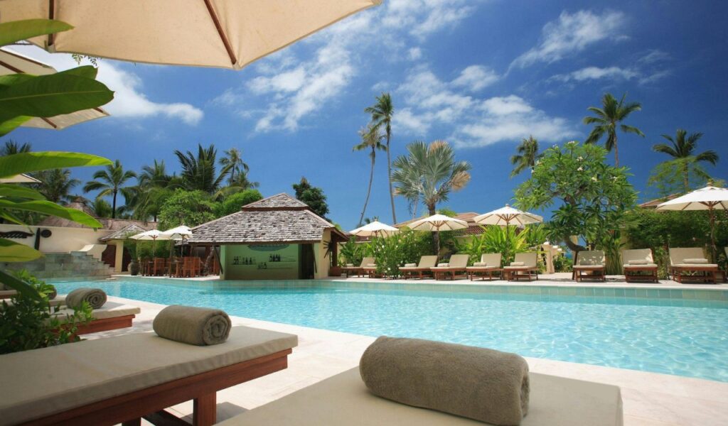 A tropical resort pool surrounded by lounge chairs, umbrellas, and lush palm trees under a clear blue sky with scattered clouds.