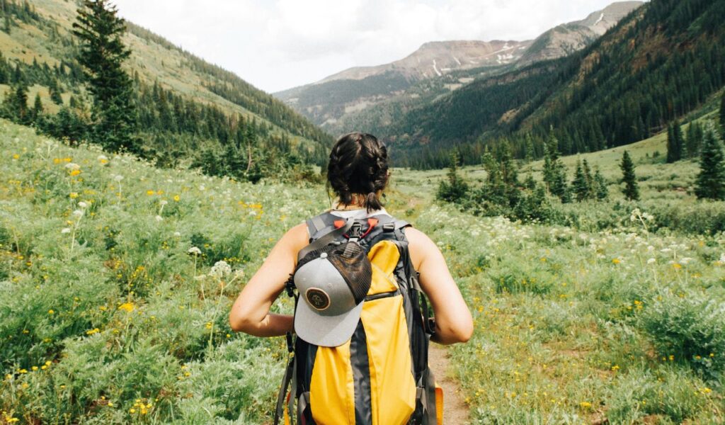A woman wearing a yellow backpack walks along a narrow trail through a wildflower-filled alpine valley surrounded by tall green mountains.