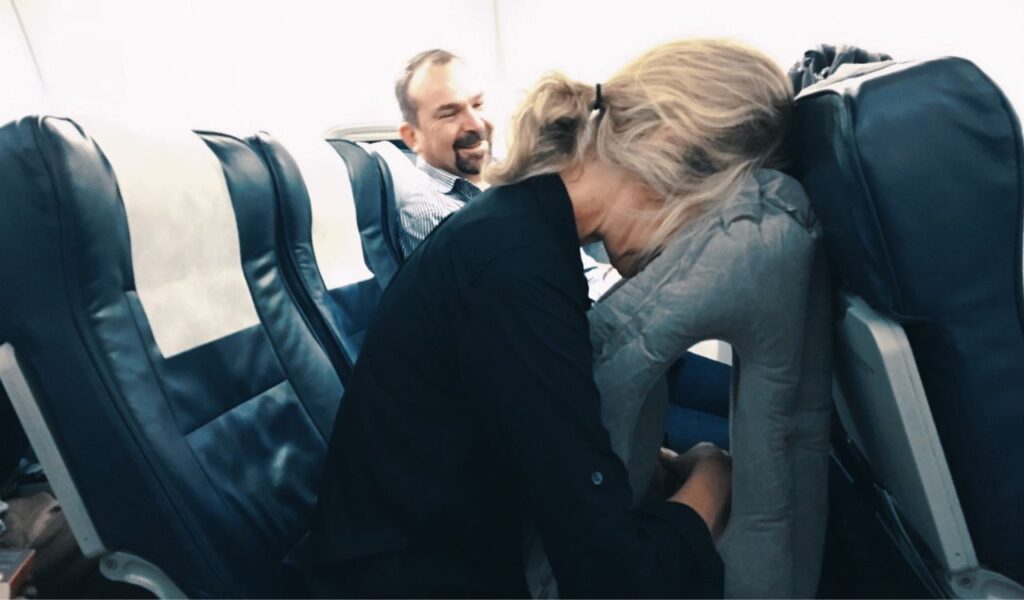 Woman using inflatable face-down travel pillow on airplane while man in next seat smiles.