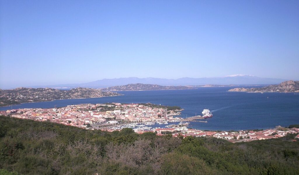 Coastal view of Palau, Sardinia with harbor, red-roofed town, blue sea, and distant mountains under clear sky.