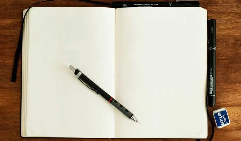  Open blank notebook on a wooden desk with a mechanical pencil, pens, and a Staedtler eraser placed around it.