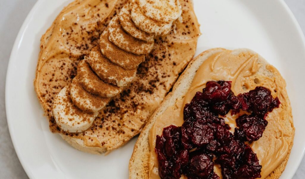 Two slices of bread topped with creamy peanut butter—one with banana slices and cinnamon, the other with a cherry or berry preserve—served on a white plate.