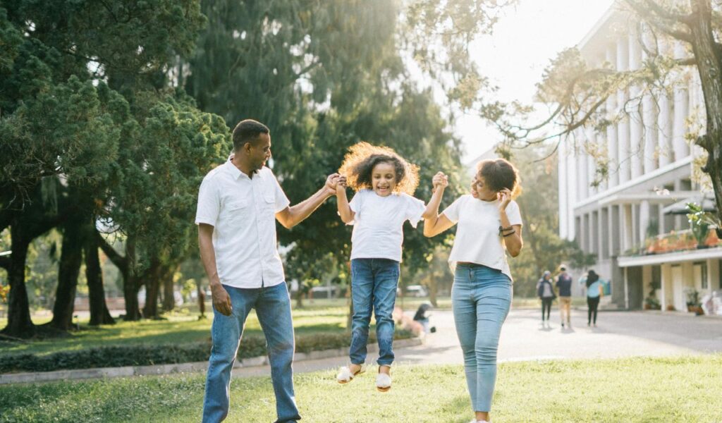 A joyful family walks through a sunny park as parents lift their smiling child by the arms, capturing a playful and carefree moment together