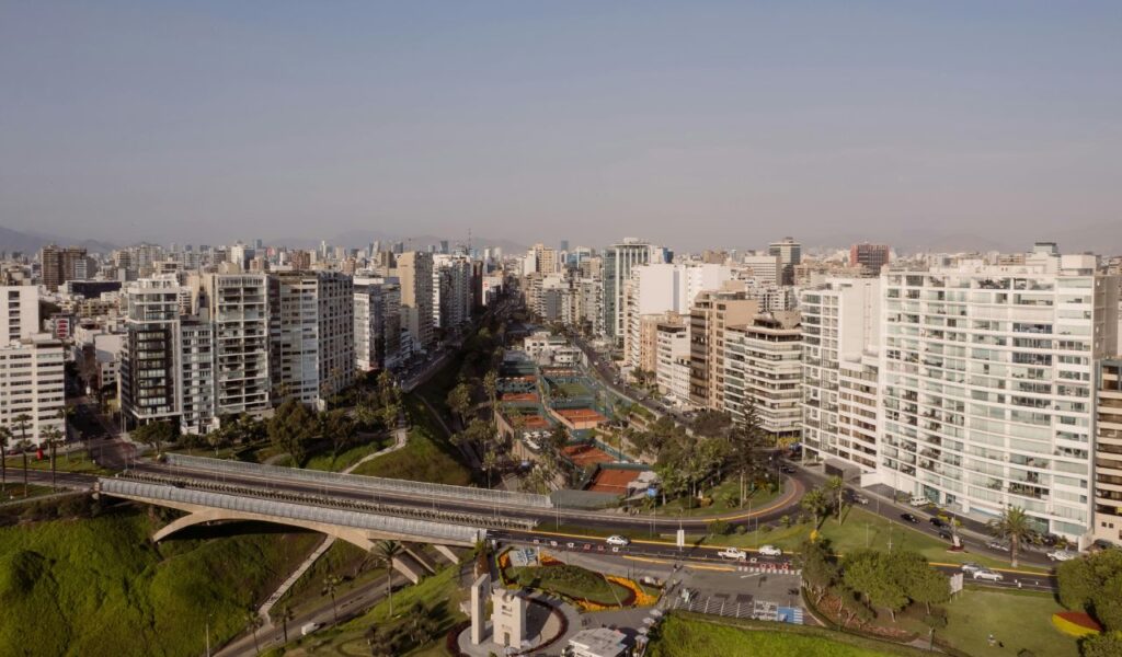 A panoramic view of Lima, Peru, showcasing modern high-rise buildings, green spaces, and a bridge, with mountains visible in the hazy background.