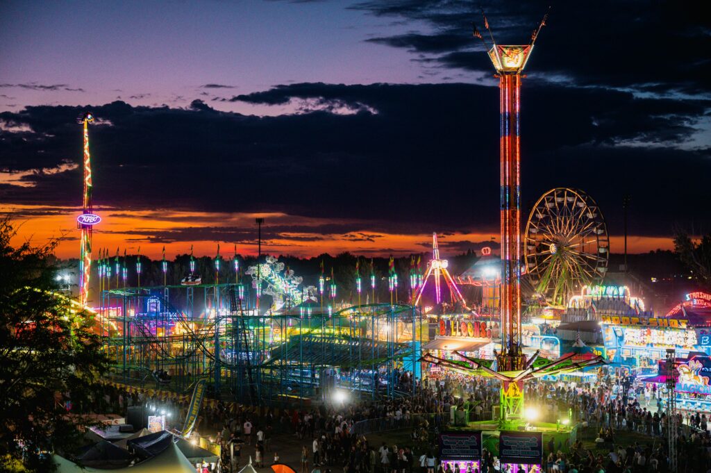 Crowds enjoying a brightly lit amusement park at dusk, with colorful rides, a Ferris wheel, and roller coasters glowing under a vivid purple-orange sunset sky.