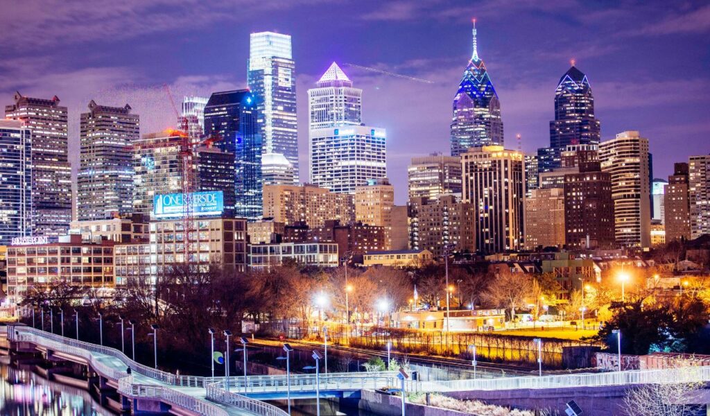 Philadelphia skyline at night with illuminated skyscrapers, glowing streetlights, and a curved pedestrian bridge over the Schuylkill River.