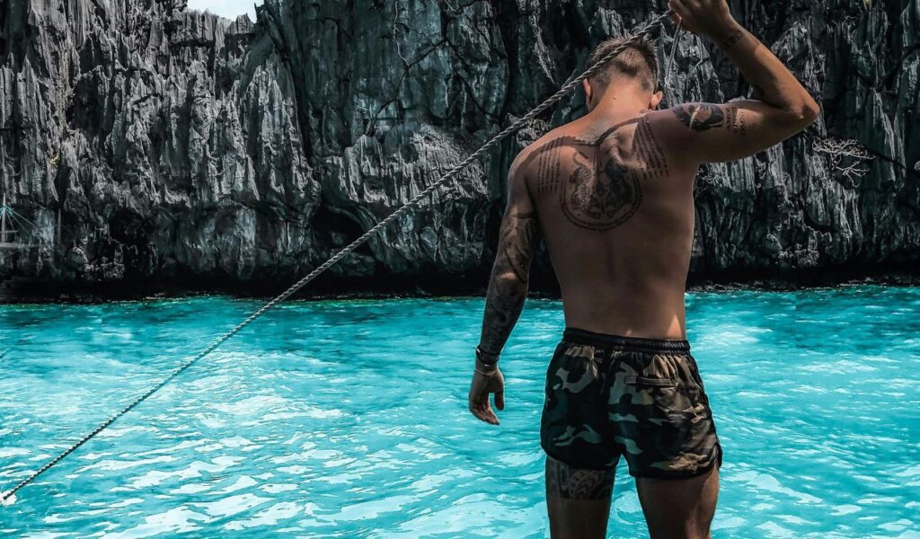 Tattooed solo traveler standing by turquoise water and limestone cliffs in the Philippines.