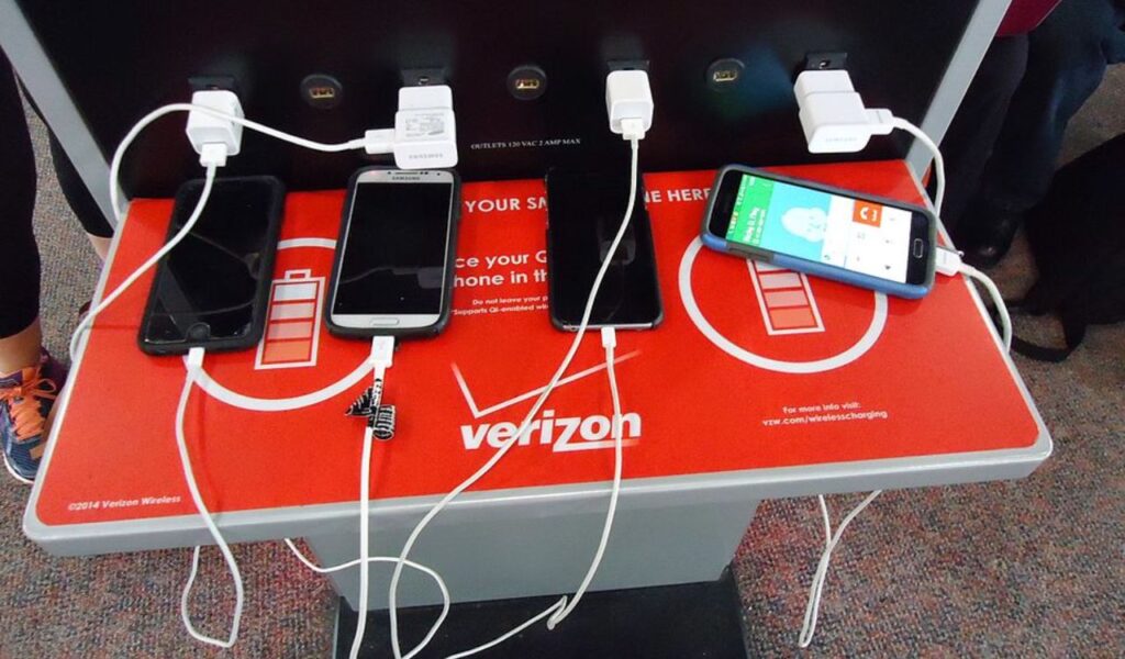 Multiple phones charging at a Verizon charging station in an airport, with cables plugged into charging ports.