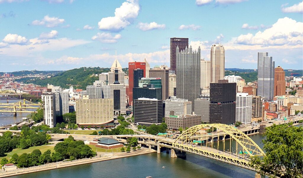 Downtown Pittsburgh skyline with iconic yellow bridges and rivers, seen on a sunny day with green hills in the background.