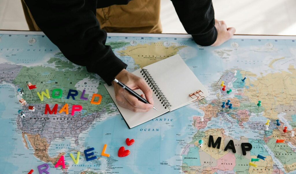 Person planning a trip by writing in a notebook placed on a world map, with colorful letters spelling "WORLD MAP" and "TRAVEL" along with pushpins marking locations across Europe and Africa.