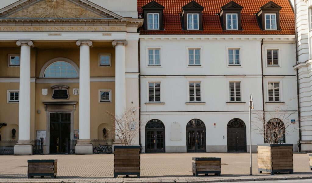 A neoclassical building with large white columns stands next to a white three-story townhouse with red roof dormers in a European city.