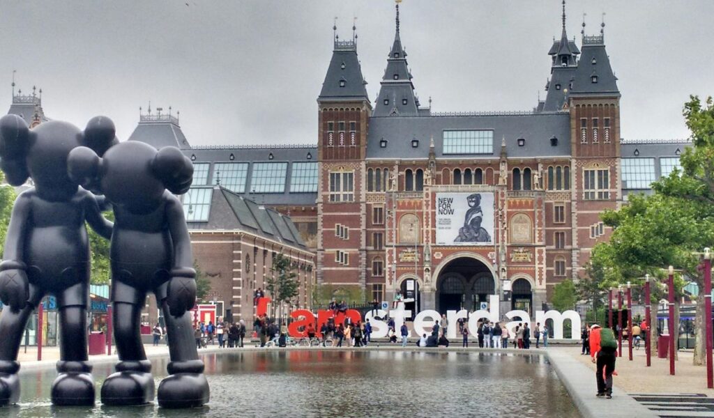 The Rijksmuseum in Amsterdam with its grand architecture, reflecting pool, "I amsterdam" sign, and crowds of visitors on a cloudy day.