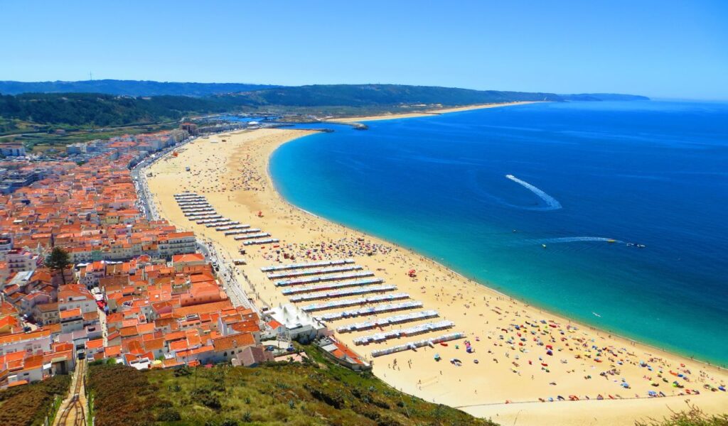 Aerial view of a vibrant beach in Portugal with golden sand, turquoise waters, and colorful umbrellas next to a charming coastal town with red-tiled roofs.