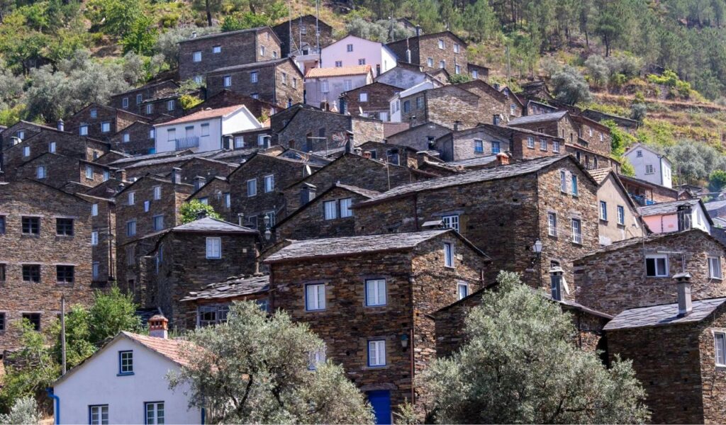 Stone houses with slate roofs densely packed on a steep hillside in a Portuguese mountain village, surrounded by trees and greenery.