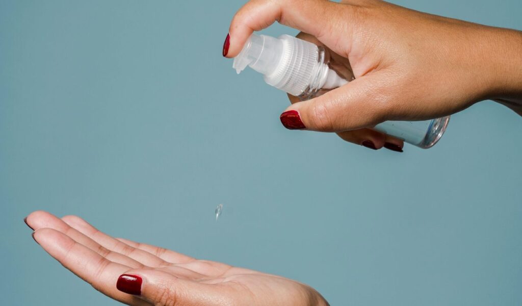 Close-up of a woman’s hands using a small spray bottle to apply moisturizer or hydrating mist against a blue background.
