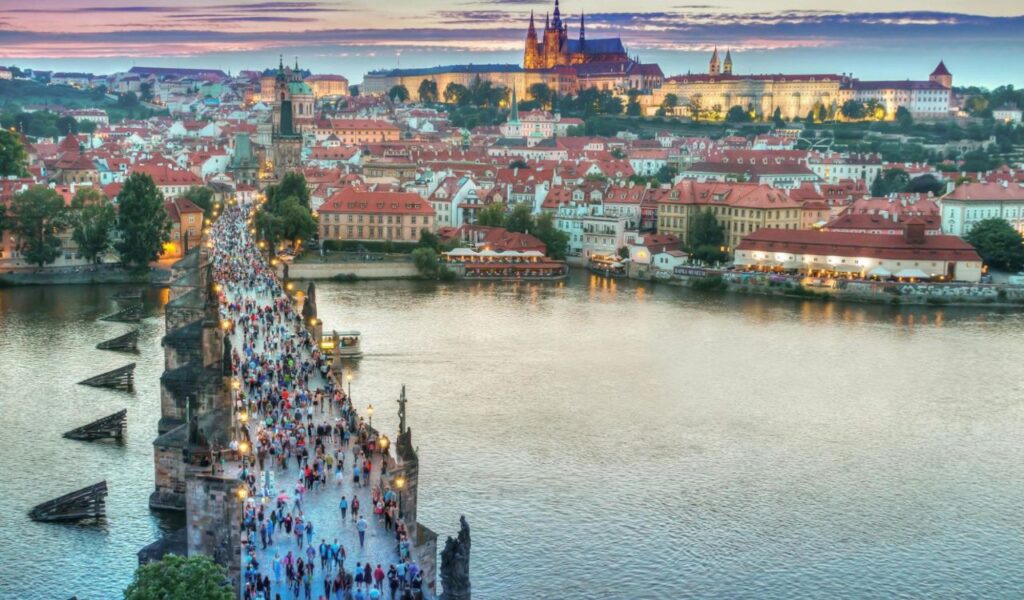 Crowds walking across Charles Bridge in Prague with Prague Castle lit up at dusk in the background.