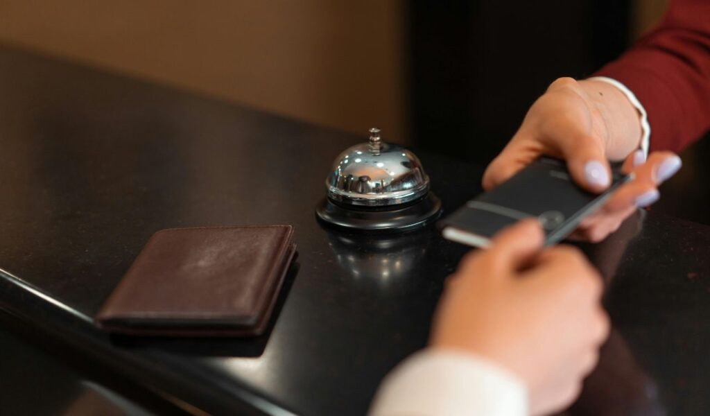 Guest making a payment at hotel front desk using a smartphone, with wallet and bell nearby implying hidden charges.