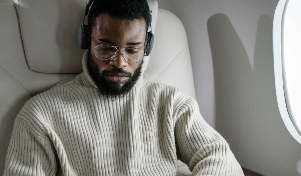 A man wearing headphones and glasses sits in an airplane seat, focused on his tablet or phone. He appears relaxed and immersed in entertainment during the flight.