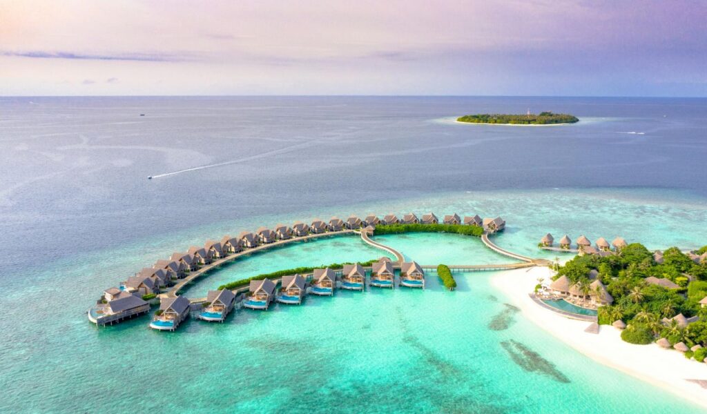Aerial view of a luxury resort on turquoise waters, featuring overwater villas arranged in a circular pattern and a white sandy beach surrounded by lush greenery.