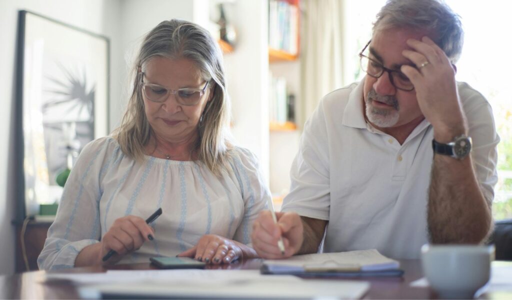 An older couple planning their trip, one using a smartphone and the other writing notes, sitting at a table in a cozy home setting.