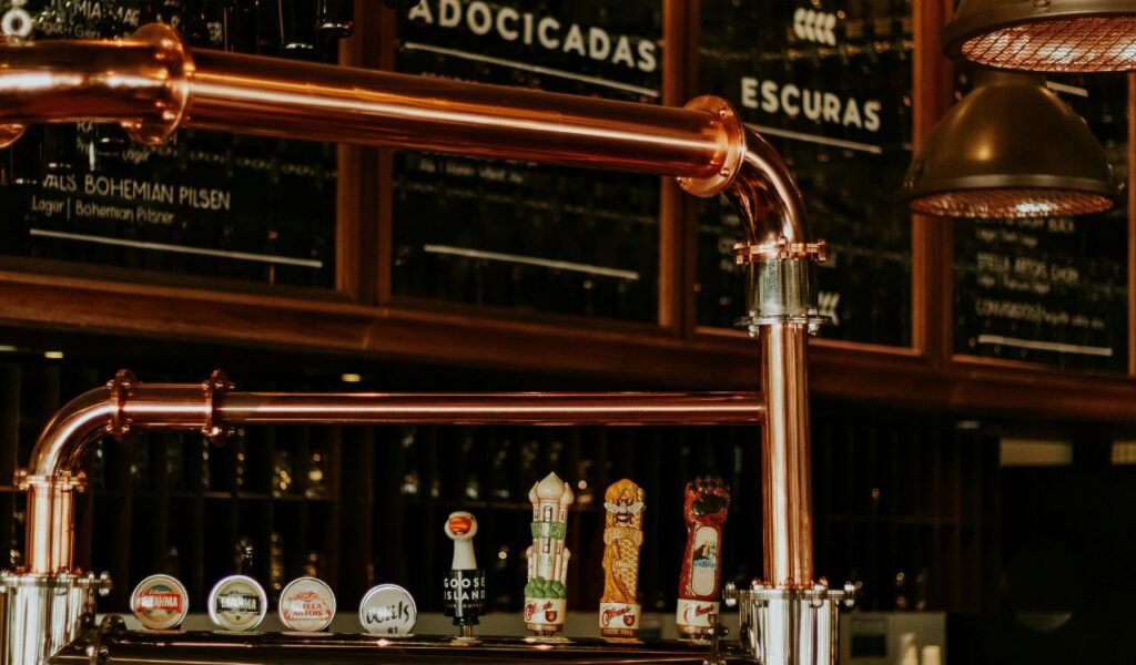 Beer taps in a Czech pub with polished copper pipes and various draft beer handles, set against a backdrop of menu boards and hanging glasses.