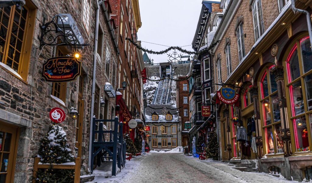 Snow-covered cobblestone street lined with decorated shops and Christmas trees in Old Quebec City, with the funicular railway visible at the end of the charming alley.