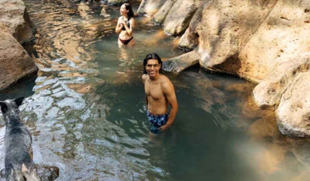 Man and woman relaxing in rocky hot spring pool with dog nearby.