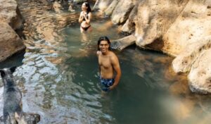 Man and woman relaxing in rocky hot spring pool with dog nearby.